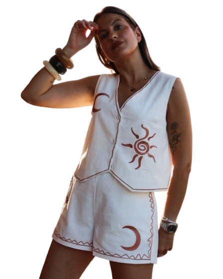 Woman wearing white linen outfit with sun and moon embroidery, posing with hand to forehead.